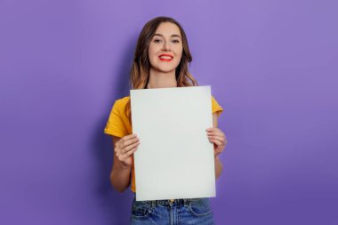 Beyaz kadın gülümsüyor ve elinde boş bir poster var sarı tişört giyiyor stüdyoda eflatun arkaplanından izole edilmiş. Tasarım için model