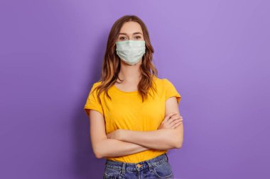 Tıbbi maske takan genç bir kadın kollarını katlayıp leylak arka planda izole olarak duruyor.