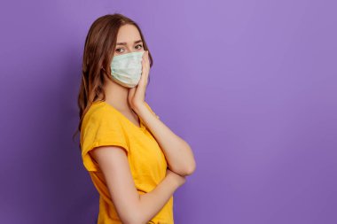 Tıbbi maske takan genç bir kadın, elini yüzünü leylak arka planında soyutlanmış bir şekilde tutuyor. Boşluğu kopyala