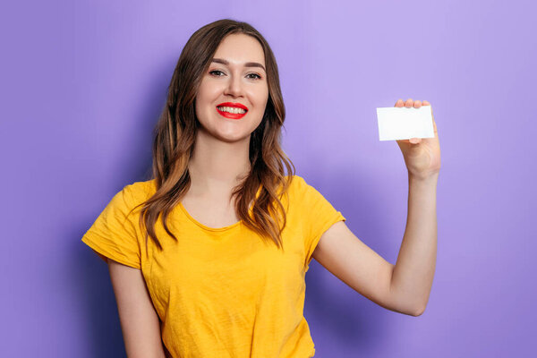 Молодая белая женщина держит визитку в руке и улыбается, одетая в желтую футболку на сиреневом фоне в студии. Кредит онлайн. Mockup