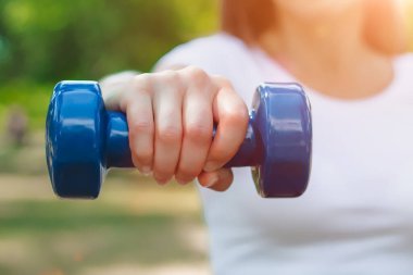 Spor eğitimi konsepti. Kadın el, arka planda mavi bir dambıl tutuyor. Kız doğada fitness ile meşgul.