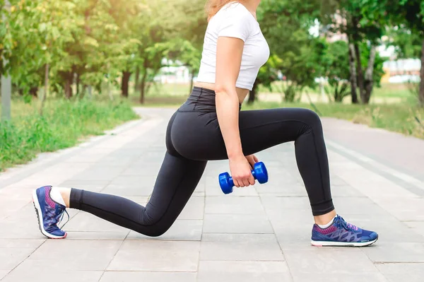 Sports girl in a white T-shirt and black leggings doing exercises with dumbbells in the park. Exercise lunge, the concept of a healthy lifestyle and proper nutrition.