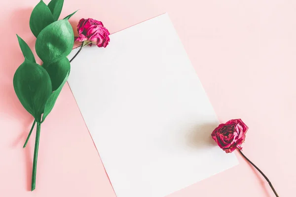 White sheet of paper on a pink background with green leaves and a dry pink rose top view mockup. Floral background for lettering with copy space.