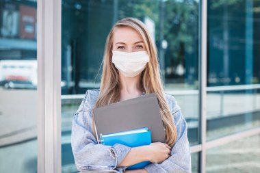 Coronavirus karantina salgını sırasında sırt çantası ve kitaplarla dışarıda poz veren doktor maskeli genç bir kızın portresi.