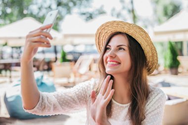 Hasır şapkalı neşeli kız görüntülü sohbete gelir ve sahilde oturur. Mutlu beyaz kadın telefonda selfie çekiyor.