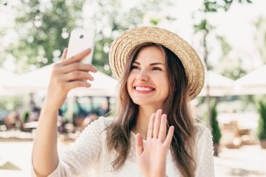 Video çağrısı konsepti. Hasır şapkalı neşeli kız görüntülü sohbete gelir ve sahilde oturur. Mutlu beyaz kadın telefonda selfie çekiyor.