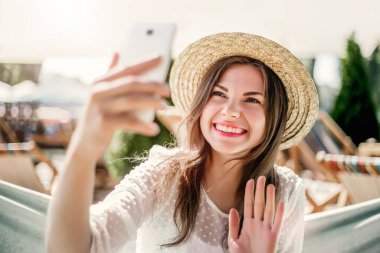 Hasır şapkalı, gülümseyen, telefonu tutan, selfie çeken, video konferans görüşmesi yapan, telefon ekranına el sallayan ve hamakta oturan mutlu ve şık bir genç kadın.