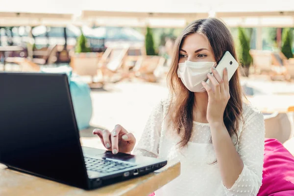 Distance work. A woman wearing a medical mask sits on the beach with a laptop at the table in a cafe and calls on a mobile phone, looking for a vacancy for a new job during the coronavirus quarantine