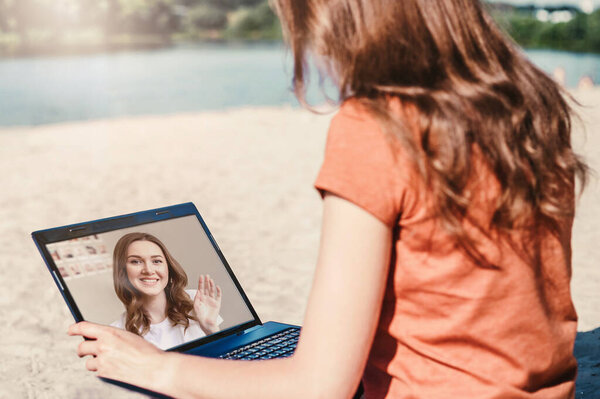 Two girls talking by videoconference on a laptop on a webcam while sitting on the beach during the coronavirus quarantine, covid-19, video calls application