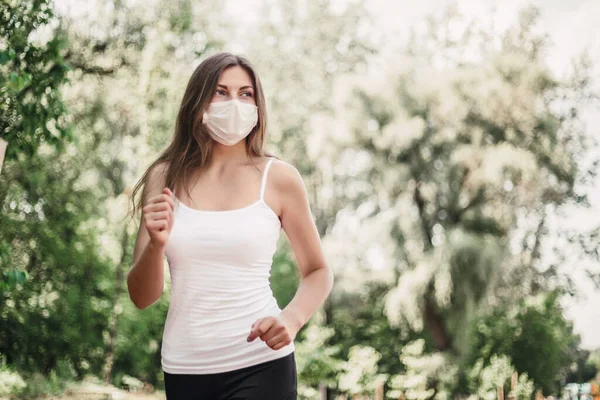 A girl wearing a protective respiratory mask runs through the park among green trees and looks at the copy space, quarantine, coronavirus 2020