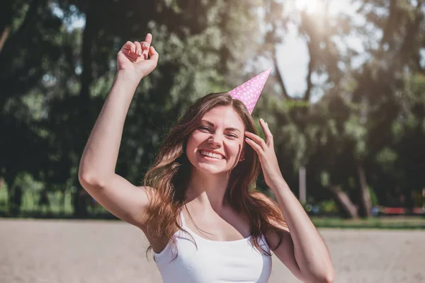 Portrait of beautiful, happy and smiling caucasian girl with a festive cone on her head dancing on the beach, birthday party alone, quarantine, coronavirus, celebration during a pandemic