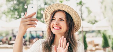 Video çağrısı konsepti. Hasır şapkalı neşeli kız görüntülü sohbete gelir ve sahilde oturur. Mutlu beyaz kadın telefonda selfie çekiyor.