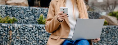 Mesafe. İş kadını genç sarışın bir kadın cep telefonu ve laptopuyla bankta oturuyor, konuşuyor ve bir ofis binasının arka planında çalışıyor.