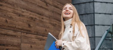 Öğrenci kız elinde defterlerin klasörlerini tutuyor gülümsüyor, modern bir üniversite binasının arka planına bakıyor. Boşluğu kopyala