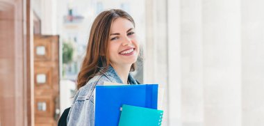 Üniversiteye karşı gülümseyen genç bir kız. Sevimli kız öğrenci klasörleri ve defterleri elinde tutuyor. Öğrenme, eğitim konsepti. Web pankartı