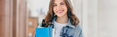 Üniversiteye karşı gülümseyen genç bir kız. Sevimli kız öğrenci klasörleri ve defterleri elinde tutuyor. Öğrenme, eğitim konsepti. Web pankartı
