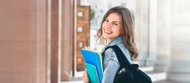 Üniversiteye karşı gülümseyen genç bir kız. Sevimli kız öğrenci klasörleri ve defterleri elinde tutuyor. Öğrenme, eğitim konsepti. Web afişi. Boşluğu kopyala