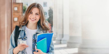 Üniversiteye karşı gülümseyen genç bir kız. Sevimli kız öğrenci klasörleri, defterleri ve cep telefonunu elinde tutuyor. Öğrenme, eğitim konsepti. Web afişi. Boşluğu kopyala