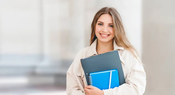 Üniversiteye karşı gülümseyen genç sarışın Ukraynalı öğrenci. Sevimli kız öğrenci klasörler ve defterler tutuyor. Beyaz arka plan. Öğrenme, eğitim konsepti. Web afişi. Boşluğu kopyala