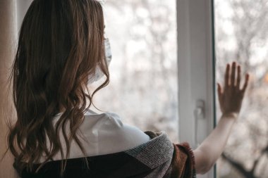 Tıbbi maskeli genç bir kadın pencereden dışarı bakar ve elini pencere camında tutar. Karantina, izolasyon, koronavirüs. Evde güvende olun.