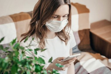 Endişeli bir kız evde karantina altında oturuyor, elinde bir cep telefonu tutuyor ve koronovirüs hakkında haberler okuyor. Uzaktan çalışan genç bir kadın, evde boş zaman, karantina.