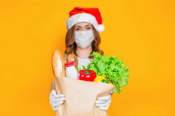 Safe food delivery for new year holidays. Courier girl in Santa Claus hat wearing a medical mask holds a paper bag with groceries, vegetables, herbs isolated on orange background