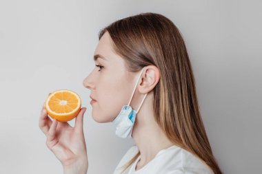 Loss of smell concept. Caucasian young woman in a medical mask sniffing an orange on a white background in the studio. profile view clipart