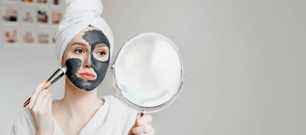 頭にタオルをつけた白いバスローブを着た若い女の子が鏡を手に持ち 問題肌 隔離隔離 ホームスパの治療のために活性炭付きの自然な黒いマスクを適用します バナー コピースペース — ストック写真