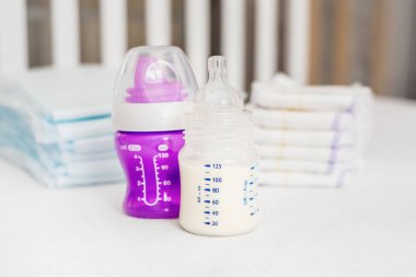 Beyaz bir yatakta bebek bulaşıkları. Tabak, kaşık, mor şişe arka planda su için beşik korkuluğu. Bebek maması. Bebek bakımı