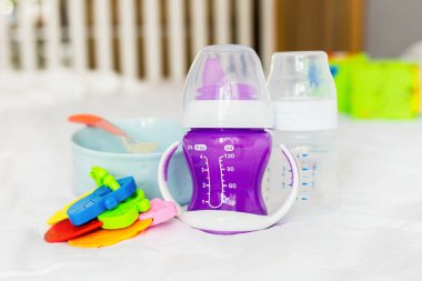 Beyaz bir yatakta bebek bulaşıkları. Tabak, kaşık, su şişesi beşik parmaklıklarının arkasında. Bebek maması. Bebek bakımı