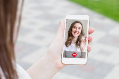 Genç, beyaz bir kadın sokakta cep telefonu tutuyor ve kız arkadaşıyla görüntülü konuşma, video görüşmeleri, konferans yapıyor. Beyaz kız cep telefonu ekranında elini sallıyor.