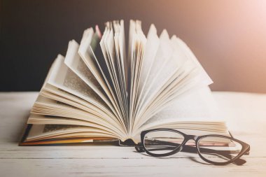 Ahşap bir masanın üzerindeki gözlüklü açık bir kitap. Arka plandaki kitaplara karşı, klasik tonlama. Edebiyat, öğrenme konsepti