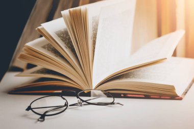 Ahşap bir masanın üzerindeki gözlüklü açık bir kitap. Arka plandaki kitaplara karşı, klasik tonlama. Edebiyat, öğrenme konsepti