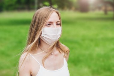Korumacı tıbbi maskeli mutlu beyaz bir kızın portresi gülüyor, gülüyor ve bahçede ağaçların ve çimenlerin üzerinde koşuyor.