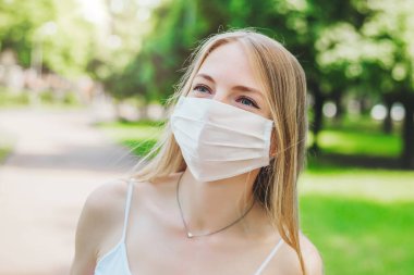 Beyaz tıbbi maskeli mutlu sarışın bir kızın portresi. Şehirde bir parkta gülümsüyor ve yürüyor. Yaklaş.