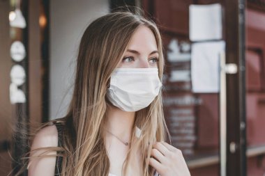 Kafkasyalı sarışın bir kızın tıbbi maske taktığı ve şehrin sokaklarında bir kafenin ön cephesinde duran portresini yakından çek.