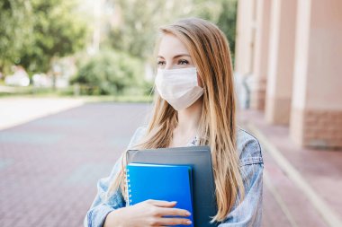 Medikal koruyucu maske takan üzgün beyaz kız üniversite ve üniversitenin önünde duruyor. Not defterlerini tutuyor ve Coronavirus salgını sırasında gözlerini kaçırıyor.