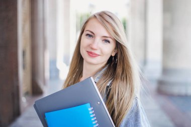 Elinde defter, klasörler ve üniversitenin arka planında gülümseyen bir kız öğrenci.