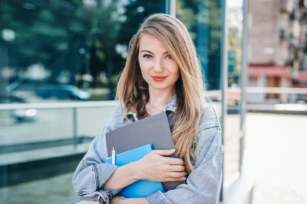 A young IT company employee stands in front of a high tech building with glass windows and holds folders and a notebook in her hands