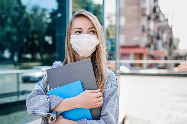 A young IT company employee wears in a medical protective mask stands in front of a high tech building with glass windows and holds folders and a notebook in her hands. copy space