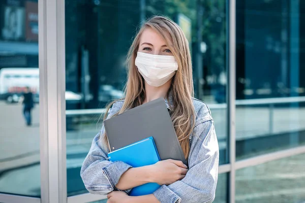 A girl in a medical mask stands under the building of a business center, holds a folder in her hands and waits for a job interview during a coronavirus quarantine