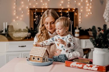 Küçük oğluyla birlikte bir anne, yeni yıl süsleri ve çelenkleriyle mutfakta bir masada oturuyor. Coronavirus karantinası sırasında zencefilli kurabiye evi yapıyorlar. DIY el sanatları, çocuklarla vakit geçirme