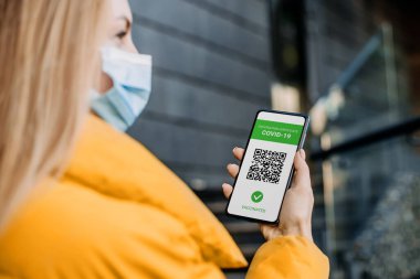 Tıbbi maske takan öğrenci Coronavirus aşısı ile ilgili bir uygulama içeren bir cep telefonu ekranı gösteriyor.