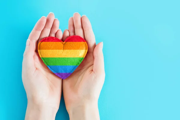 Female hands are holding a rainbow heart symbol of the LGBT community flag on a blue background, greeting card, background for poster, flyer, banner, copy space. LGBT background. Female hands painted in LGBT flag