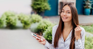 Kafkasyalı genç bir kadın cep telefonu kullanıyor gülümsüyor ve caddedeki kameraya bakıyor ağaçların arka planına ve bir kafe cephesine karşı. Mesafeli iş. İnternet afişi. Boşluğu kopyala