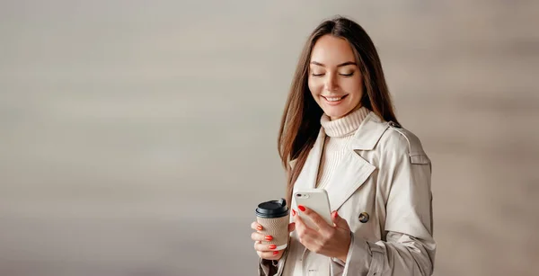 A student girl wearing in a trench coat uses a mobile phone, looks at the screen, smiles against the backdrop of the facade of an office building. web banner. copy space