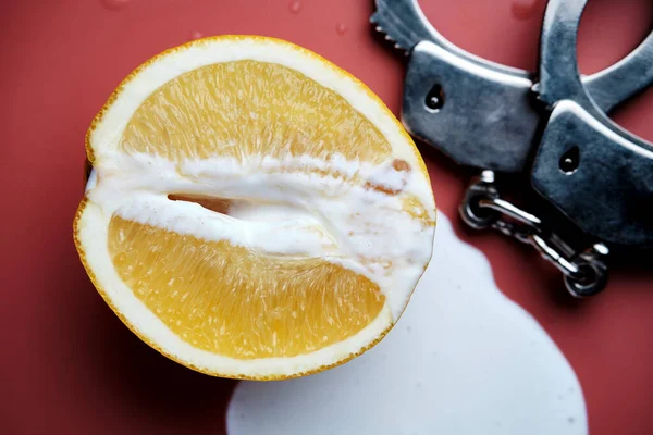 Half an orange, leaking yogurt and a fragment of handcuffs on a pink background. Erotic concept. A symbolic image of a vagina and sperm. Unprotected sex. Close-up. Selective focus