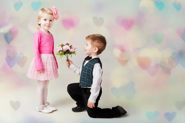 Der kleine Junge kniet auf einem Knie und schenkt dem Mädchen Blumen. Herzen bokeh Hintergrund. Es ist das erste Valentinstagsgeschenk. Romantisches Geschenk — Stockfoto