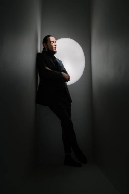 Sakin, üzgün, yakışıklı, uzun saçlı ve sakallı bir adam duvara doğru çaprazlama eğiliyor ve düşünceli bir şekilde yere bakıyor. Moda pozu. Sahne ışığı ile koyu renkler dikey bileşimi