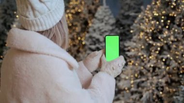 Cep telefonu yeşil ekran şablonu. Kışın giyinmiş bir kadın karla kaplı ormanda ağaçlarla süslenmiş bir şekilde tutuyor. Sihirli kış tatili anları. Renkli anahtar ekranlı video görüntüleri 